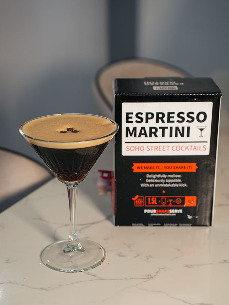 A prepared Espresso Martini cocktail in a coupe glass with a thick layer of crema and a coffee bean on top, placed beside its black branded box labeled 'ESPRESSO MARTINI SOHO STREET COCKTAILS' on a light countertop.